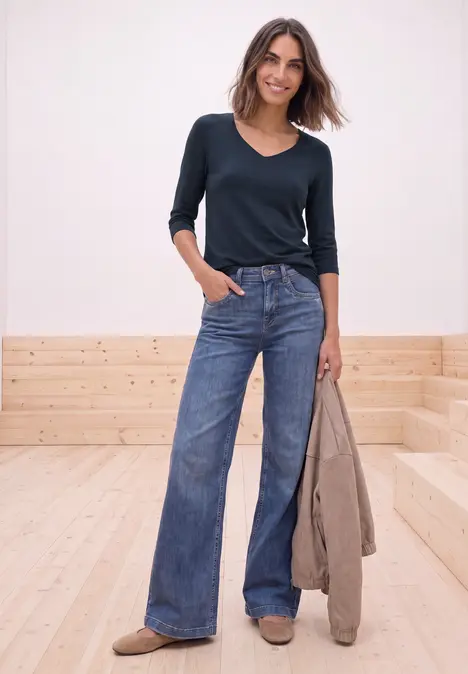 Frau in navyblauem V-Shirt und weiten Jeans, hält beige Jacke vor hellem Holz-Wand und -Boden.
