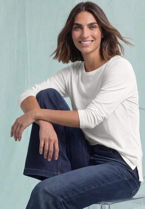 Eine lächelnde Frau im weißen Langarmshirt und Jeans sitzt entspannt auf einem Hocker.