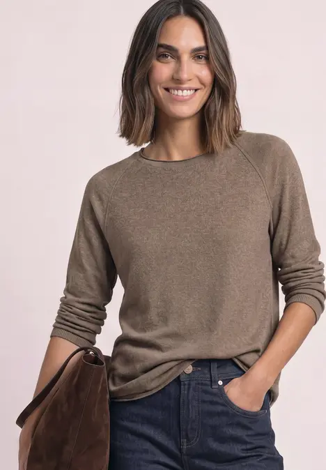 Vrouw met halflang haar draagt een bruin trui, jeans en houdt een bruine tas vast, ze lacht ontspannen.