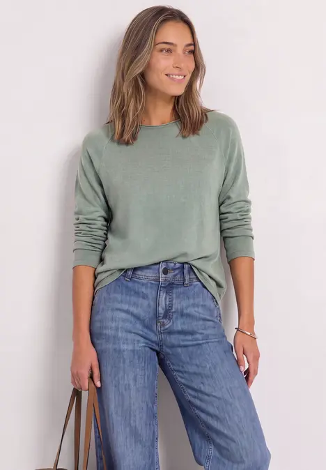 Eine lächelnde Frau in grünem Pullover und blauen Jeans hält eine Tasche.
