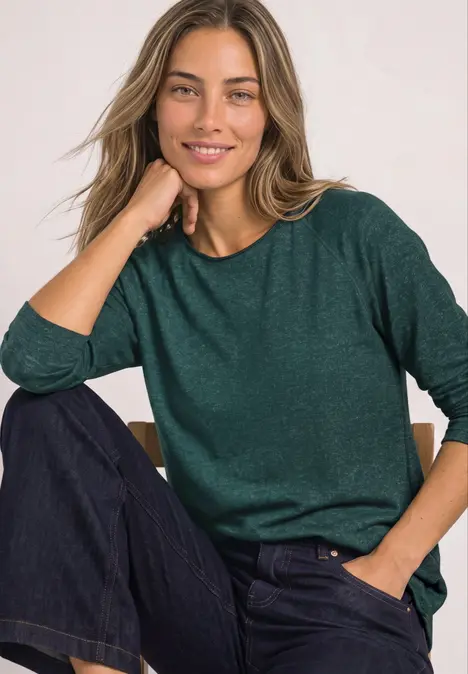 Lächelnde Frau im grünen Pullover und Jeans, sitzend und stützt das Kinn auf die Hand.