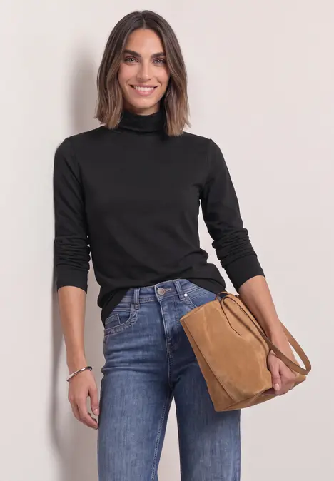 Une femme souriante en pull col roulé noir et jean, portant un sac à main marron.
