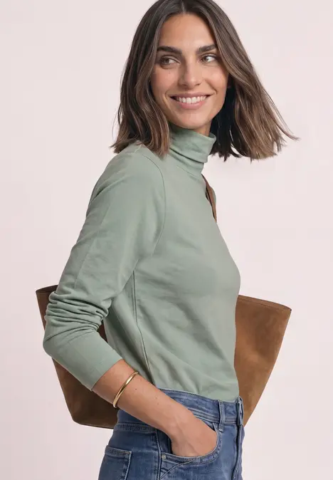 Eine lächelnde Frau in grünem Rollkragenpullover und Jeans, mit brauner Tasche über der Schulter.