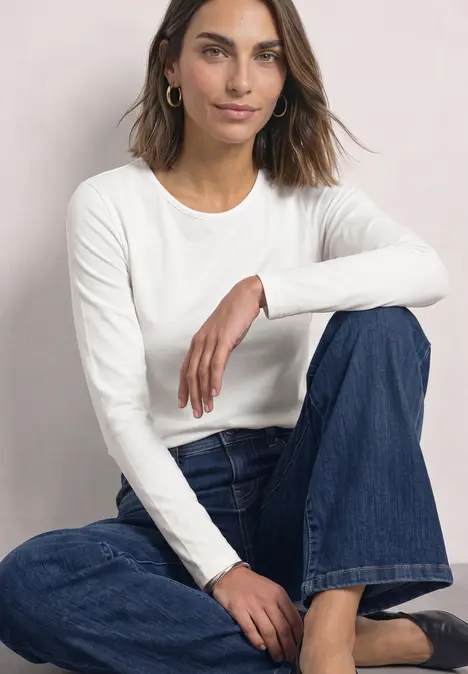 Eine Frau sitzt lässig vor hellem Hintergrund, trägt weißes Langarmshirt und blaue Jeans.
