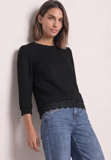 Frau in schwarzem Langarmshirt mit Spitzenbesatz und Jeans vor hellem Hintergrund.