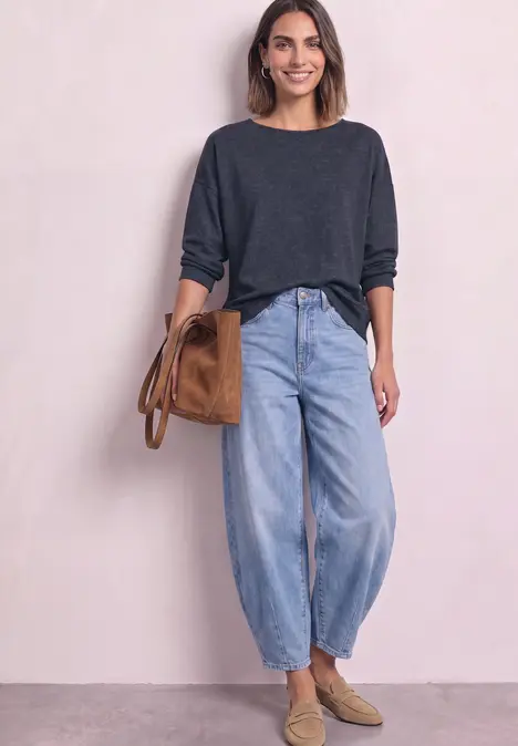Vrouw in grijze trui, losse jeans, beige loafers en een bruine tas, staand voor een lichte achtergrond.