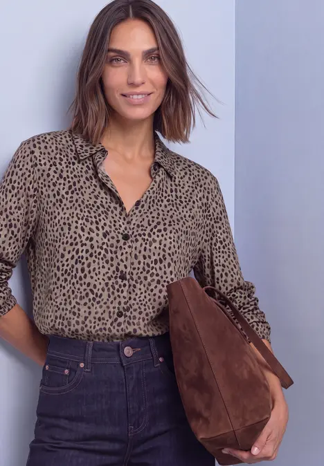 Eine Frau trägt eine beige Bluse mit schwarzen Punkten, dunkle Jeans und eine braune Wildledertasche.