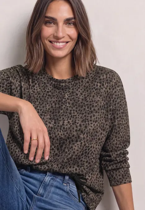 Frau mit schulterlangen braunen Haaren in graubraunem Leopardenprint-Pulli und blauen Jeans lächelt.