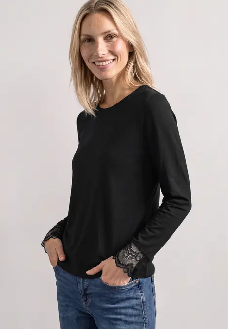 Uśmiechnięta blondynka ma na sobie czarną bluzkę z długimi rękawami i koronkowymi mankietami oraz jeansy.