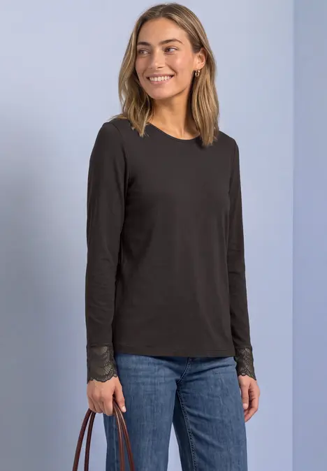 Junge Frau lächelt, trägt schwarz langärmliges Shirt, blaue Jeans und hält eine braune Tasche.