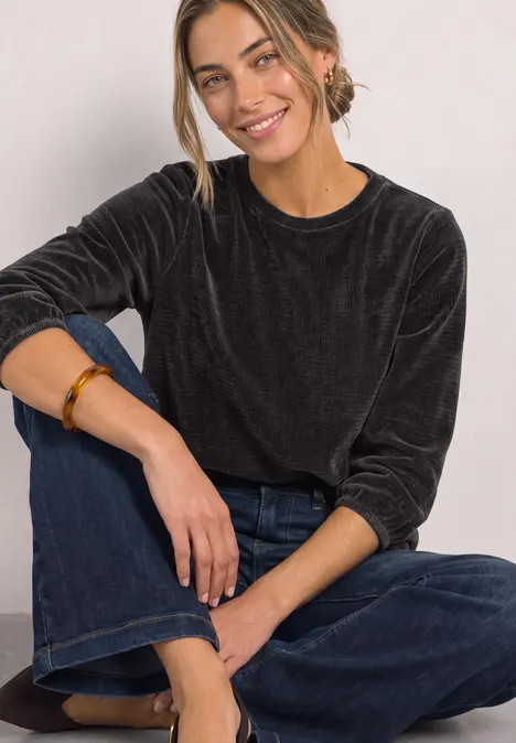Junge Frau sitzt entspannt auf dem Boden, trägt schwarzen Samt-Pullover und weite Jeans, lächelt.