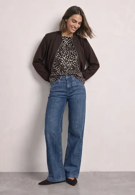 Frau in lässigem Outfit: dunkelbraune Jacke, gemustertes Oberteil und weite Jeans posiert locker vor neutralem Hintergrund.