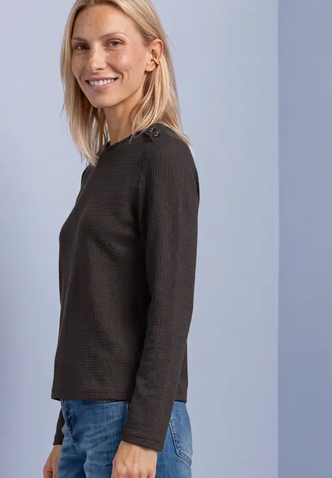 Eine blonde Frau in schwarzem Langarmshirt und blauer Jeans lächelt seitlich in die Kamera.
