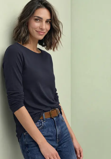 Een glimlachende vrouw in donkerblauw shirt met lange mouwen en jeans leunt tegen een lichte muur.