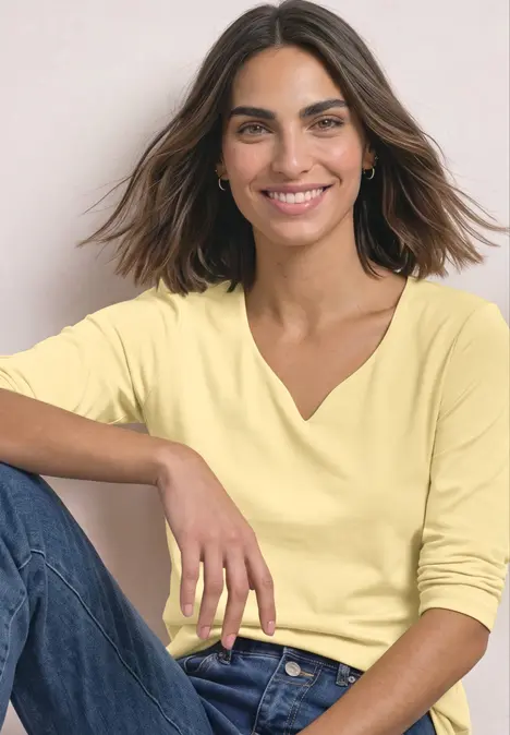 Une femme souriante porte un haut jaune et un jean, assise de manière décontractée.