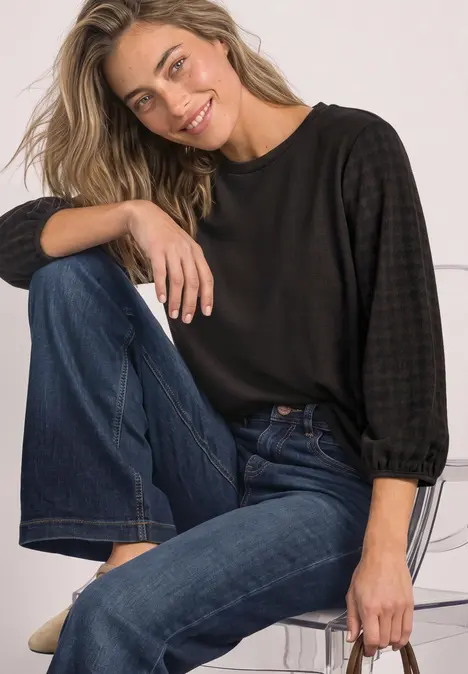 Eine lächelnde Frau sitzt entspannt auf einem Stuhl, trägt schwarzen Pullover und blaue Jeans.