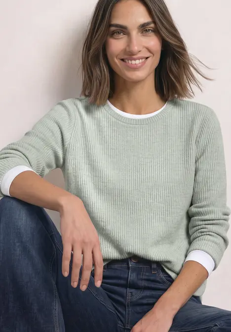 Une femme souriante porte un pull vert pâle et un jean bleu, assise devant un fond clair.