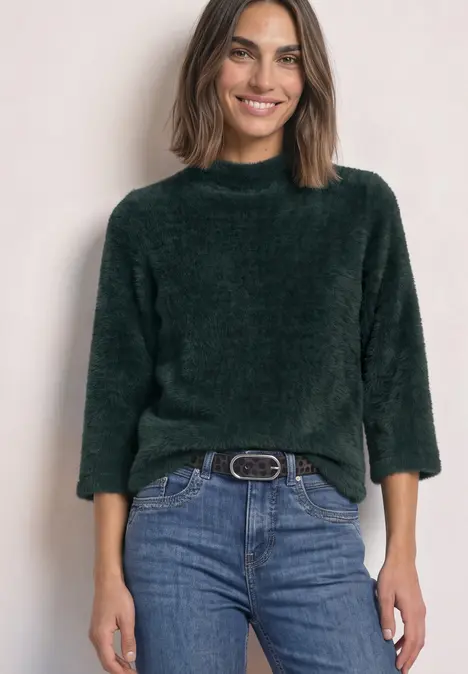 Femme portant un pull vert douillet à manches 3/4 et un jean, elle sourit.