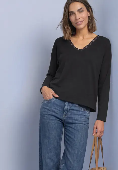 Frau in schwarzem V-Ausschnitt-Pullover und Jeans hält eine Tasche vor hellem Hintergrund.