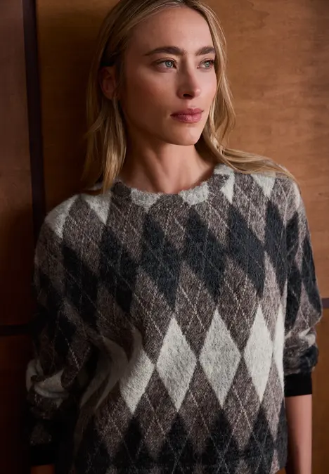 Femme portant un pull à losanges beige, marron et noir devant un mur en bois