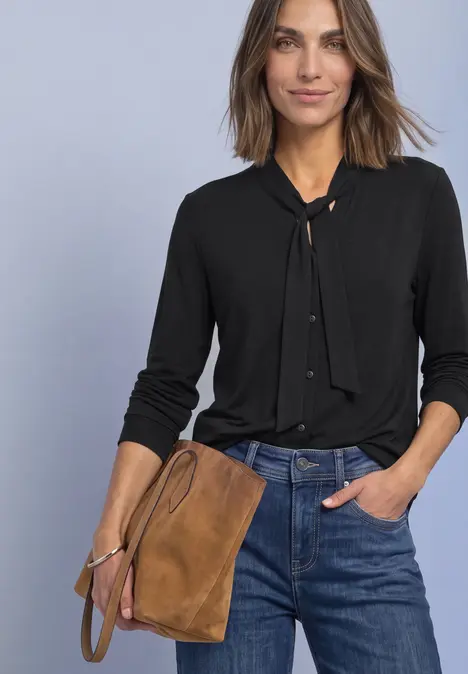 Frau trägt schwarze Bluse mit neckholder-Knoten, blaue Jeans und eine braune Wildledertasche.
