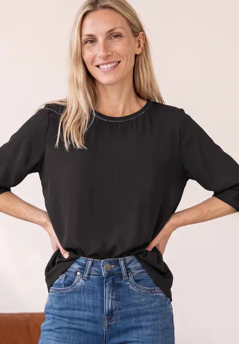 Lächelnde Frau mit schwarzem T-Shirt und Jeans, steht mit Händen in der Hüfte vor neutralem Hintergrund.