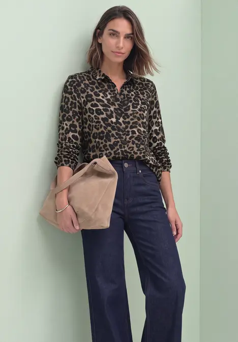 Eine Frau vor grünem Hintergrund trägt ein Leopardenmuster-Hemd, weite Jeans und hält eine beige Tasche.