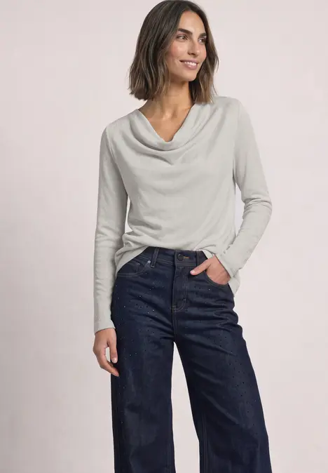 Frau trägt graues Langarmshirt mit Wasserfallausschnitt und dunkle Jeans, steht lächelnd vor hellem Hintergrund.