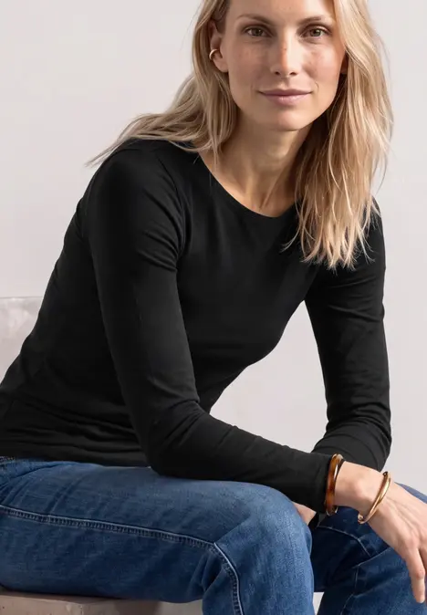 Une femme aux cheveux blonds longs, portant un haut noir et un jean bleu, est assise.