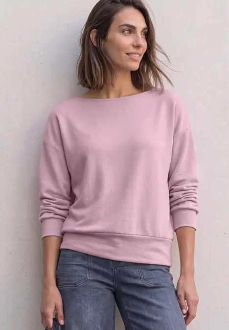 Frau steht lässig vor Wand, trägt rosa Sweatshirt und Jeans, Blick seitlich, entspannte Pose.