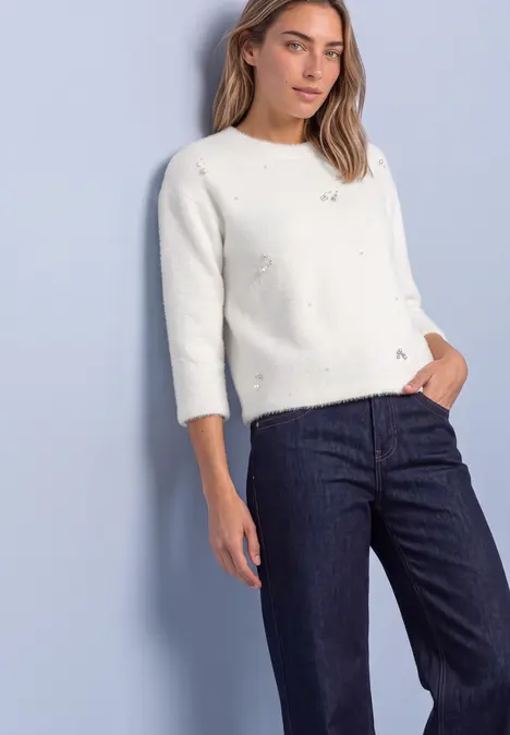 Femme en pull blanc à motifs ajourés et jean bleu foncé sur fond pastel bleu.