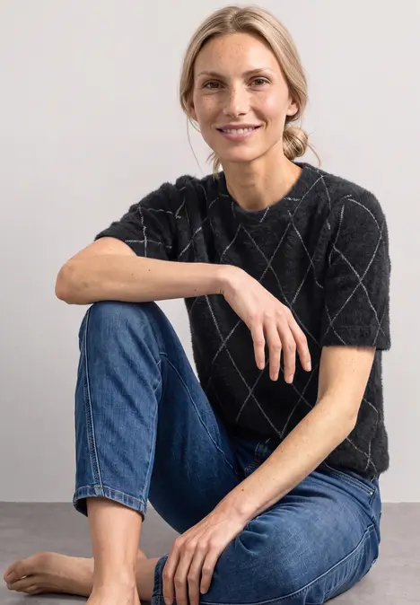 Eine Frau sitzt barfuß auf dem Boden, lächelt, trägt blaue Jeans und ein kariertes schwarzes Oberteil.