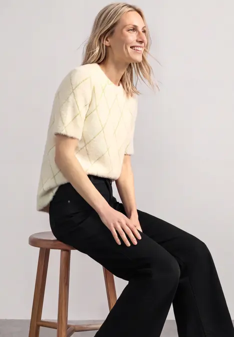 Blonde Frau sitzt auf Hocker, lächelt und trägt kurzärmligen, hellen Strickpullover mit schwarzen Hosen.