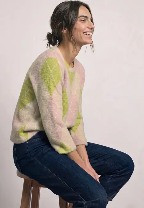 Eine lächelnde Frau sitzt seitlich auf einem Hocker und trägt Jeans und einen hellgrünen-pinken Karopullover.
