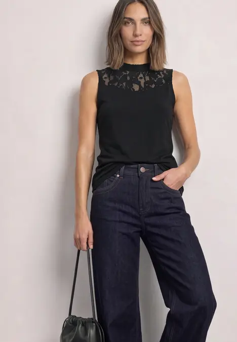 Eine Frau trägt ein ärmelloses Spitzen-Top in Schwarz, weite Jeans und hält eine schwarze Tasche.