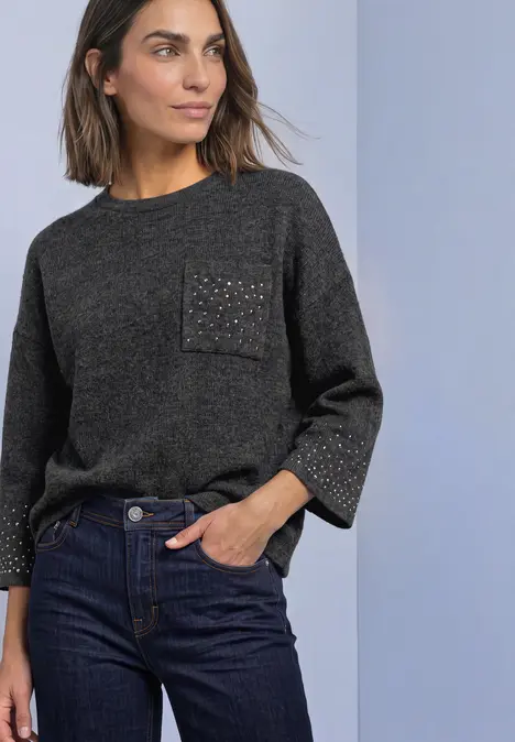 Frau in dunkelgrauem Pullover mit funkelnder Tasche und dunklen Jeans vor hellem Hintergrund.