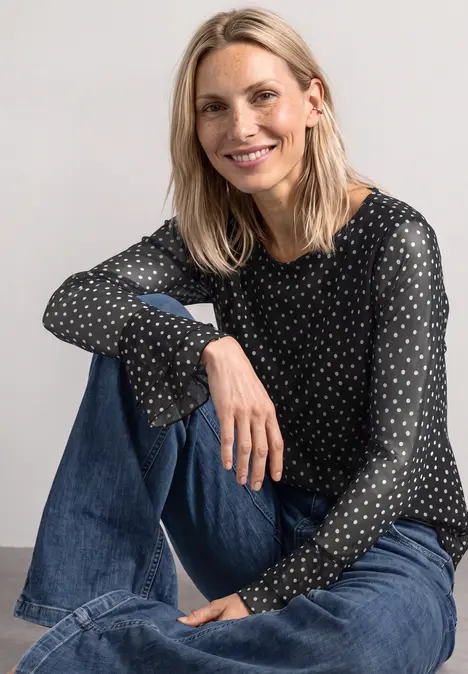 Eine lächelnde blonde Frau sitzt locker in Jeans und schwarzer Bluse mit weißen Punkten vor neutralem Hintergrund.