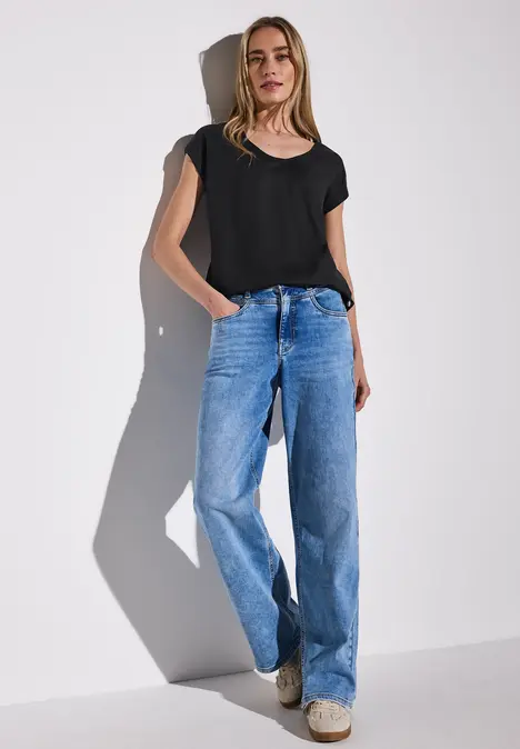 Frau mit schwarzem T-Shirt und Jeans steht vor weißer Wand.