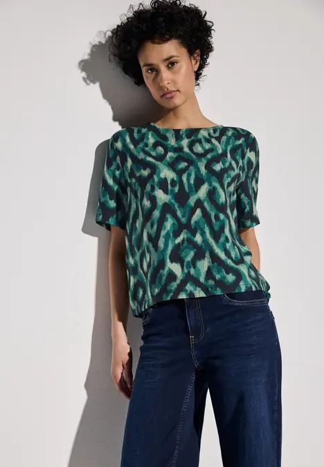 Frau mit lockigem Haar trägt ein gemustertes grünes Shirt und dunkelblaue Jeans.