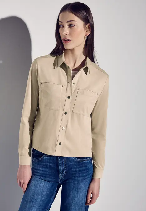 Frau mit beiger Bluse und blauer Jeans steht vor hellem Hintergrund.