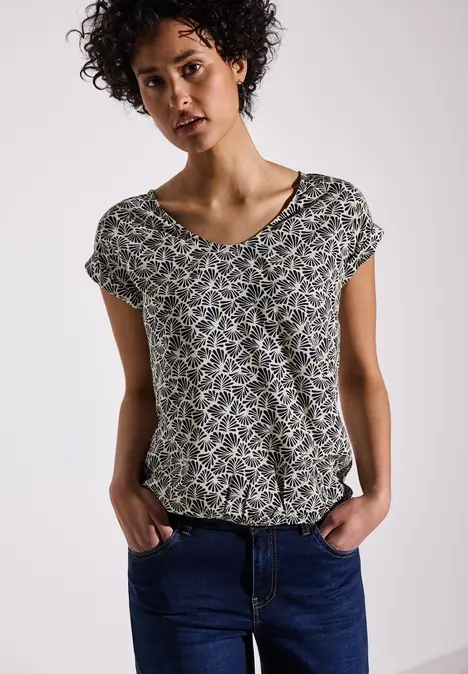 Frau mit lockigen Haaren trägt ein gemustertes Shirt und blaue Jeans.