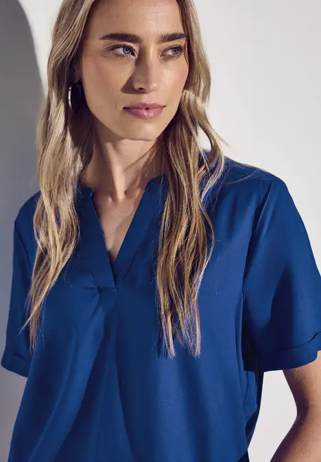 Frau mit langen blonden Haaren trägt eine blaue Bluse und blickt zur Seite.