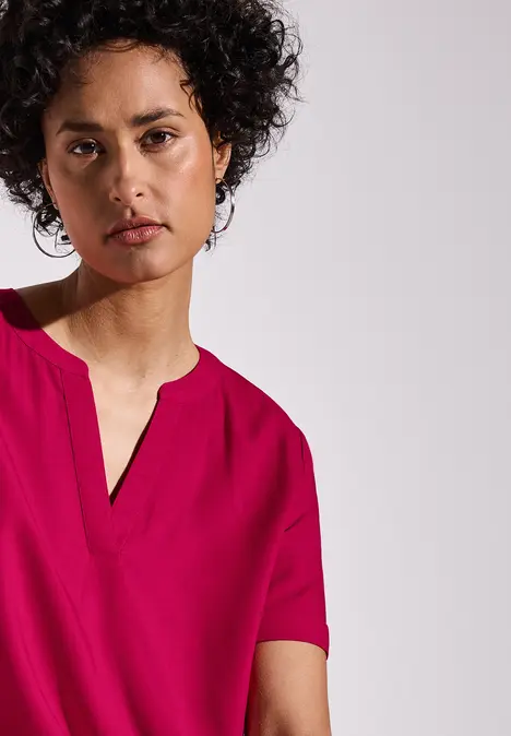 Frau mit lockigem Haar trägt ein pinkes Shirt und schaut ruhig in die Kamera.