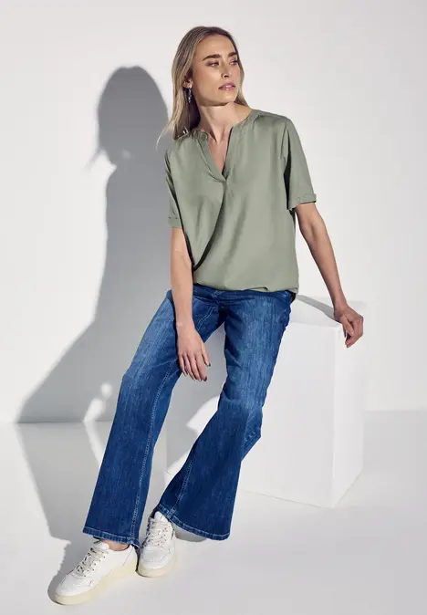 Frau mit grüner Bluse und blauen Jeans sitzt entspannt auf weißem Würfel.