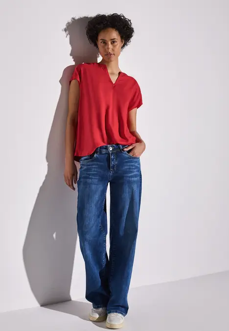 Frau mit rotem Oberteil und blauen Jeans lehnt an der Wand.