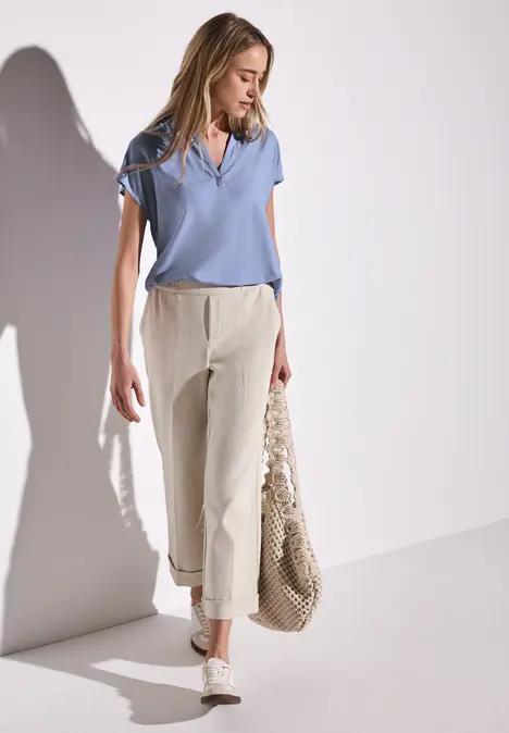 Frau mit hellblauem Shirt, heller Hose, Tasche und Sneakers geht an weißer Wand.