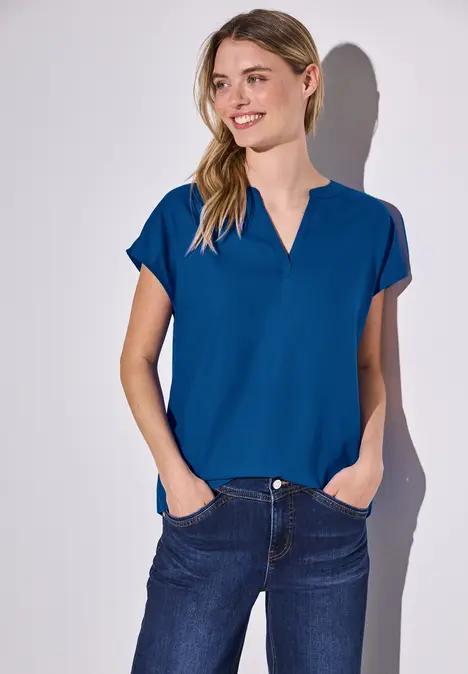 Frau in blauem Shirt und Jeans steht lächelnd vor weißer Wand.