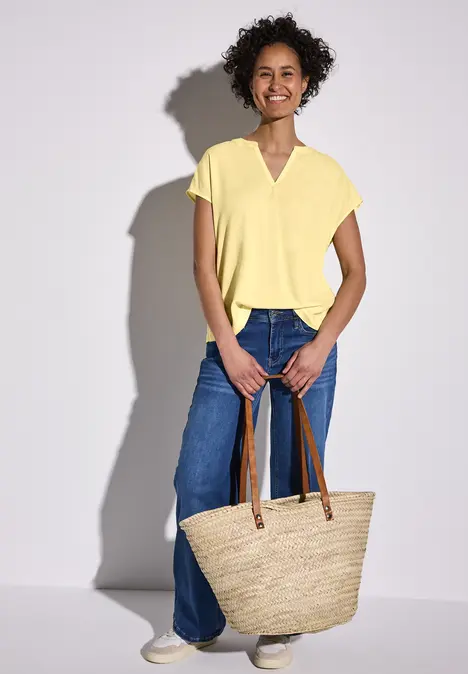 Frau mit gelbem Shirt, Jeans und großer Strohtasche lächelt.