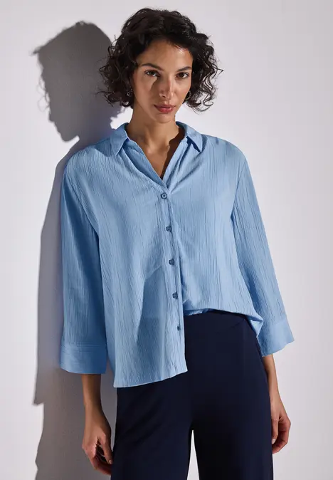 Frau mit lockigem Haar trägt eine hellblaue Bluse und dunkelblaue Hose.