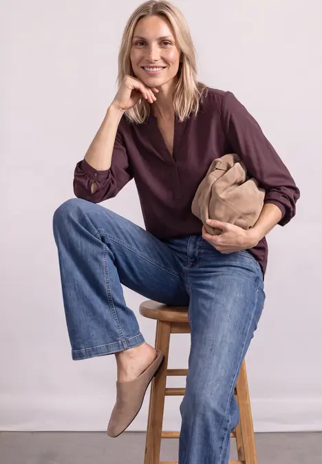 Eine lächelnde Frau mit blonden Haaren sitzt auf einem Hocker in brauner Bluse, blauen Jeans und hält eine Tasche.